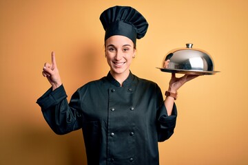 Young beautiful chef woman wearing cooker uniform and hat holding tray with dome surprised with an idea or question pointing finger with happy face, number one