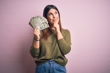 Young beautiful woman holding dollars standing over isolated pink background serious face thinking...