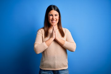 Young beautiful woman wearing casual sweater over blue background praying with hands together asking for forgiveness smiling confident.