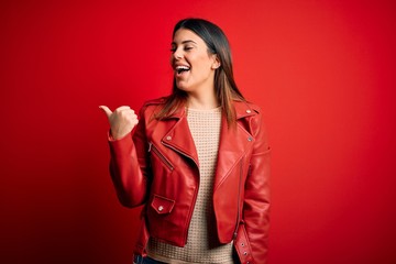 Young beautiful woman wearing red leather jacket over isolated background smiling with happy face looking and pointing to the side with thumb up.