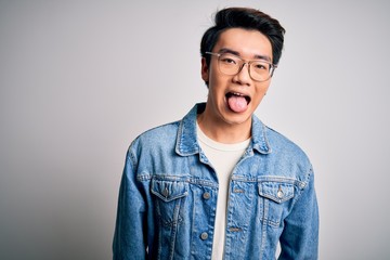 Young handsome chinese man wearing denim jacket and glasses over white background sticking tongue out happy with funny expression. Emotion concept.