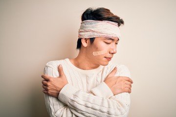 Young handsome chinese man injured for accident wearing bandage and strips on head Hugging oneself happy and positive, smiling confident. Self love and self care