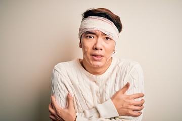 Young handsome chinese man injured for accident wearing bandage and strips on head shaking and freezing for winter cold with sad and shock expression on face