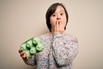 Young down syndrome woman holding cardboard egg cup from fresh healthy eggs cover mouth with hand...