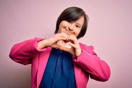 Young down syndrome business woman over pink background smiling in love doing heart symbol shape with hands. Romantic concept.