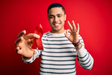 Young brazilian man holding red hiv ribbon standing over isolated background doing ok sign with fingers, excellent symbol - obrazy, fototapety, plakaty