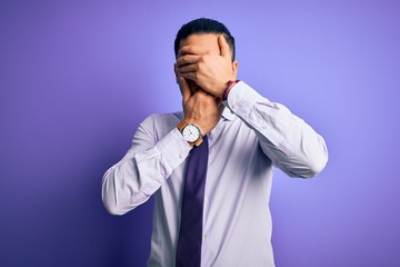 Young brazilian businessman wearing elegant tie standing over isolated purple background Covering eyes and mouth with hands, surprised and shocked. Hiding emotion