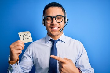 Young brazilian call center agent man holding reminder paper with yes message very happy pointing with hand and finger