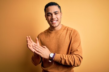 Young brazilian man wearing casual sweater standing over isolated yellow background clapping and applauding happy and joyful, smiling proud hands together