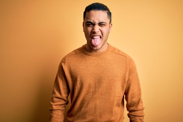 Young brazilian man wearing casual sweater standing over isolated yellow background sticking tongue out happy with funny expression. Emotion concept.
