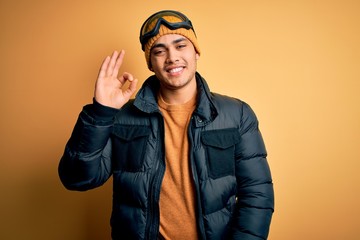 Young brazilian skier man wearing snow sportswear and ski goggles over yellow background smiling positive doing ok sign with hand and fingers. Successful expression.