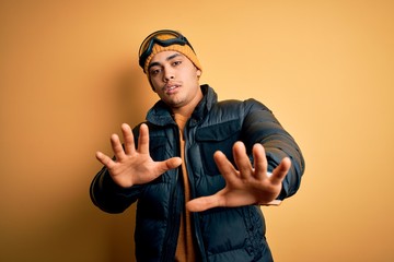 Young brazilian skier man wearing snow sportswear and ski goggles over yellow background doing stop gesture with hands palms, angry and frustration expression