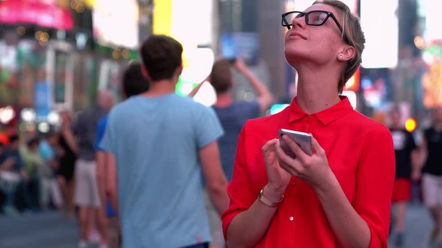 Millennial traveller in glasses with cute smile on face standing at metropolitan crowd square and looking around while thinking on content text for creating publication via cellphone gadget 
