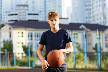 Cute smiling boy in blue t shirt plays basketball on city playground. Active teen enjoying outdoor game with orange ball. Hobby, active lifestyle, sport for kids.