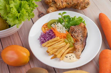 Fish steak with french fries, kiwi, lettuce, carrots, tomatoes, and cabbage in a white dish.
