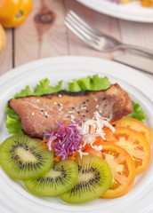 Fish steak with french fries, kiwi, lettuce, carrots, tomatoes, and cabbage in a white dish.