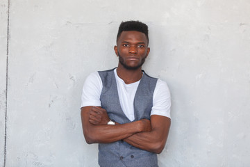 Young handsome african black man posing with crossed arms in grey vest and trousers against a gray concrete wall