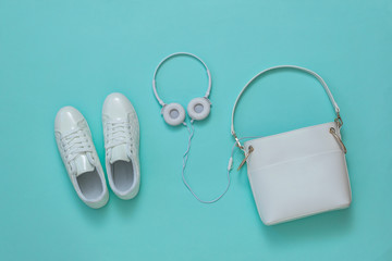 White women's bag, white headphones and white sneakers on a light blue background. Flat lay.