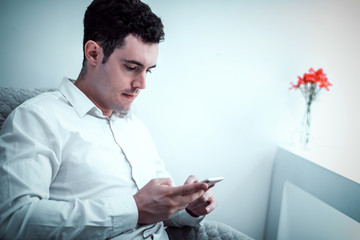 cinematic tone of young Caucasian man using smartphone or cell phone and sitting alone on chair in white room