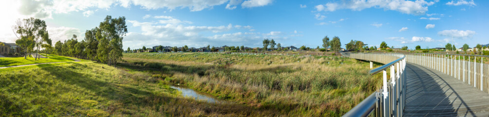 Naklejka premium Panoramic view of Skeleton Waterholes Creek with a wooden boardwalk leads to some suburban houses in distance. Truganina, Melbourne, VIC Australia.