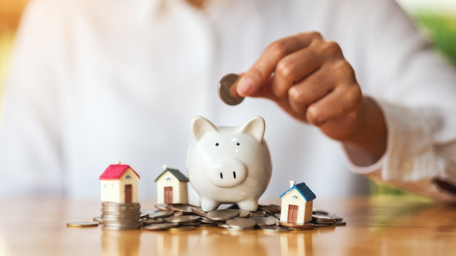 A Woman Putting Coins Into Piggy Bank With House Models On Pile Of Coins For Saving Money Concept