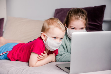 Two children brother and sister lie on the couch in medical masks using laptop, home education, stay at home.