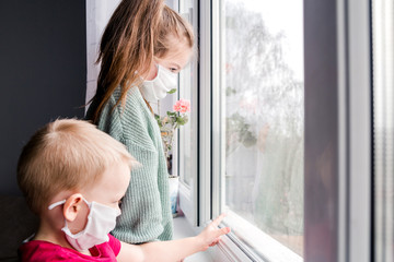 Cute Children Boy and Girl in medical Mask stay Home on Quarantine. Children play Home. Entertainment for the Children during quarantine. Self isolation. Children are looking out the window.