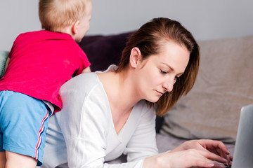 While young focused mother freelancer doing remote work or surfing internet lying on couch type on lap computer her active  kids  indulge, fooling around and stop her from working