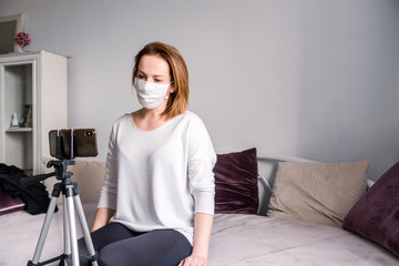 A woman in a white medical mask looks at the phone, which stands on a tripod. Video calling
