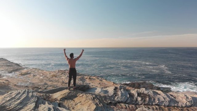 Aerial Drone Circles Athletic Man Celebrating on Ocean Cliffs Edge at Sunrise