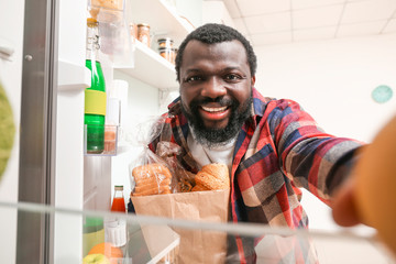 African-American man unpacking fresh products from market in kitchen, view from inside of fridge