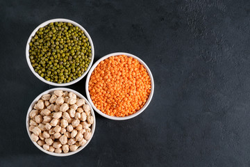 Three cups of legumes on black background - mung beans, chickpea and red lentil. Horizontal arrangement, copy space