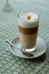 Glass with latte coffee on a saucer with a spoon. A table in a street cafe. White borders of milk and brown edges of coffee are visible in a glass. Clear day. The city of Tbilisi, Georgia.