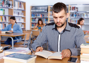 Portrait of young bearded man sitting in public library and reading books