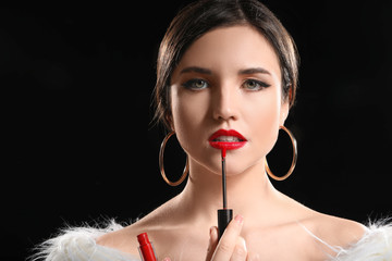 Beautiful young woman applying lipstick against dark background