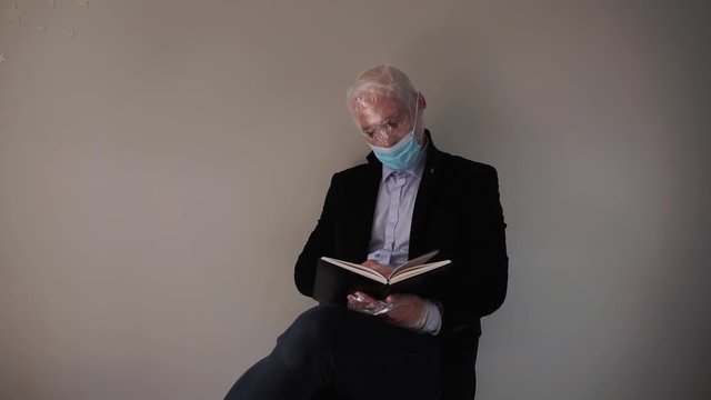 Man dressed in black jacket in protective mask and with face fully rolled up with plastic foil because of over reaction regarding corona virus situation is reading a book 