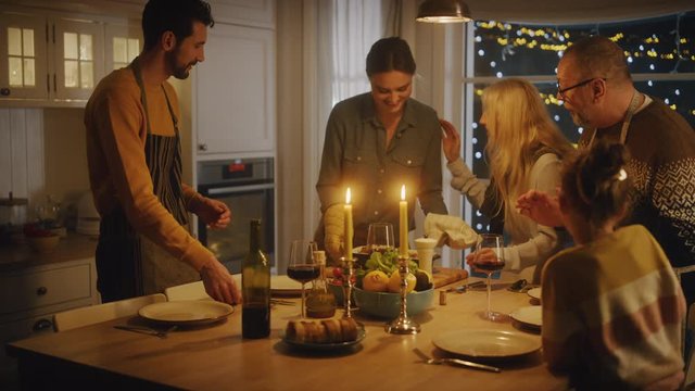 Happy Family of Celebrating together, Sitting at the Table with Delicious Dinner. Young Mother Takes Casserole out of Oven, Father Pours Wine, Daugther Listens to Grandparents. Everybody Admires Dish