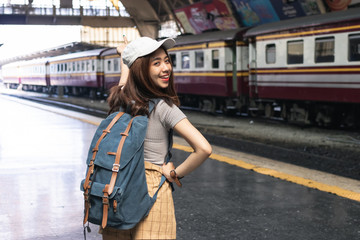 Fototapeta premium Smiling young Asian backpacker female standing at train station. Summer travel lifestyle concept.