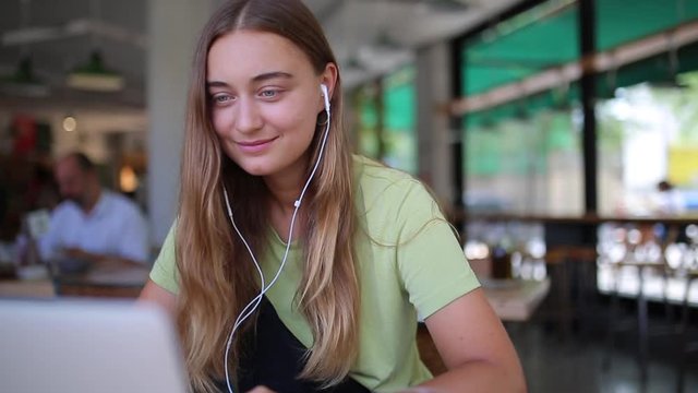 Pretty young happy millennial white female uses headphones and smiles. Freelance woman enjoys public workspace, study online e-learning, checking e-mail, news feed or works on project