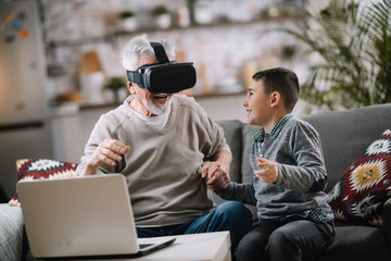 Grandson and his grandpa playing with VR at home on sofa. Grandfather and a little boy having fun at home.