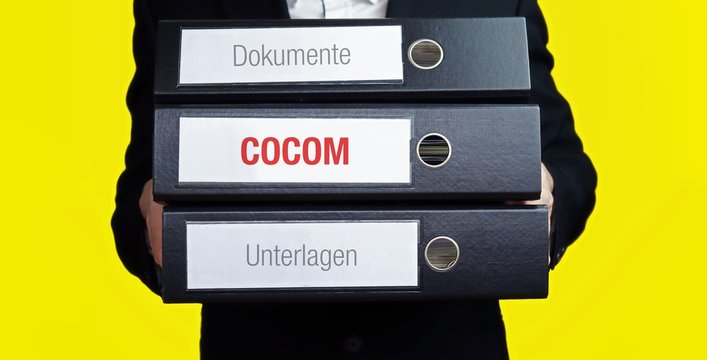COCOM &ndash; Finanzen/Wirtschaft. Mann tr&auml;gt einen Stapel Aktenordner. Ein Ordner hat die Beschriftung COCOM. Symbol f&uuml;r Business, Statistik