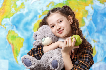 Cute teen girl with a teddy bear in her hands eats a green apple on a background of the world map