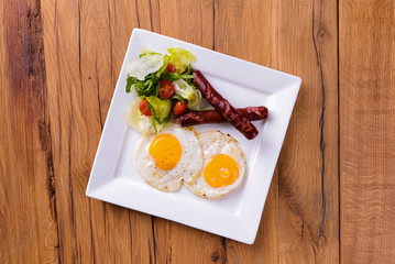 Breakfast with bacon, fried egg and tomatoes on served on white plate over light rustic wooden table.
