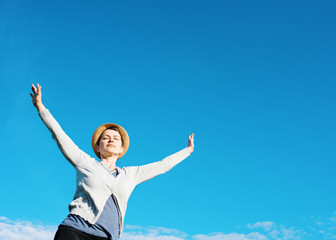 Happy woman wearing grey top, straw hat,  raised her hands high against blu sky