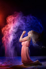 Beautiful woman with long brunette hair in pink dress with colored lights and flour around. Portrait of girl and female model posing in photoshoot with flour at studio with black background