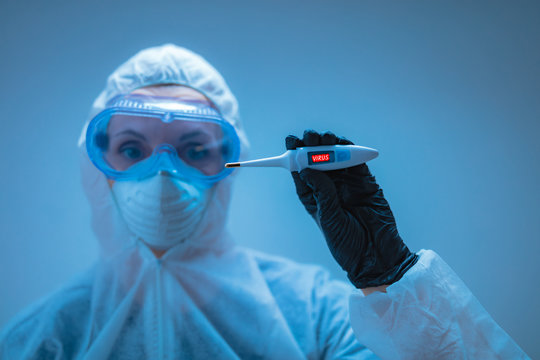 Medical Doctor, Scientist In Full Body Suit For Prevention From Viruses And Diseases Holding A Thermometer For Body Temperature.