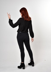 Portrait of a pretty girl with red hair wearing black jeans, boots and a blouse.  full length standing pose on a studio background, with back to the camera.
