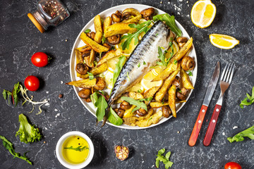Baked mackerel with lemon, baked potatoes and mushrooms on a plate on black stone background.