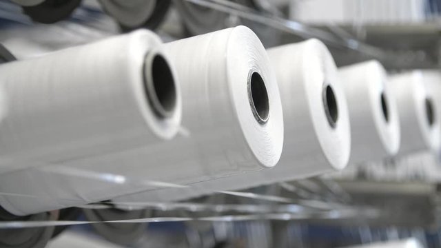 Impressive closeup of straight long of white reels revolting around and having knitting spokes producing white plastic sacks for agriculture on modern plant.