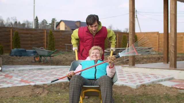 A young man is trying to ride his elderly mother in a wheelbarrow, but she is too heavy. He cannot move the barrow. Mom with a rake in her hands sits in a wheelbarrow and laughs